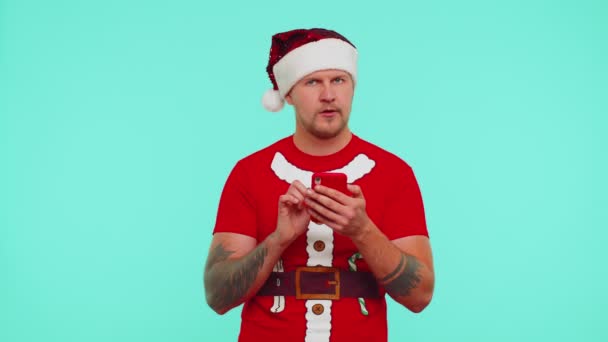 赤いクリスマスTシャツの男使用携帯電話のタイピングブラウジング損失驚きの突然の宝くじ結果 — ストック動画