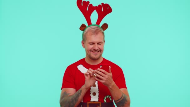 Homem de t-shirt de Natal usando telefone celular digitando novo post na web, compras on-line, mostrar polegares para cima — Vídeo de Stock