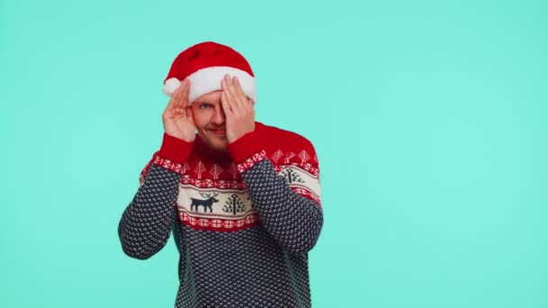 Man in sweater Santa Christmas hat fooling around having closing eyes with hand and spying through — Stock Video