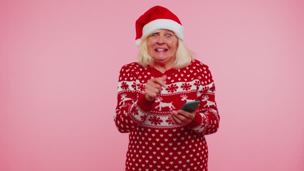 Nenek yang mengenakan sweater Natal, yang tampak seperti smartphone. Menampilkan kegembiraan yang tulus dan memenangkan keberuntungan. — Stok Video
