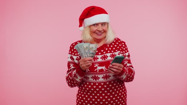 Різдвяна бабуся дивиться смартфон щиро радісно виграє успіх отримуючи гроші в Інтернеті — стокове відео