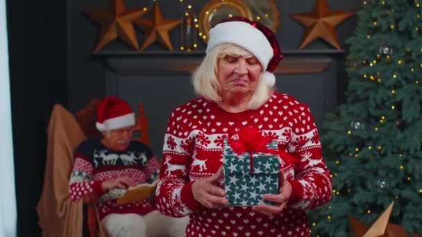 Зріла бабуся в святковому светрі представляє різдвяну подарункову коробку, посміхаючись, дивлячись на камеру — стокове відео