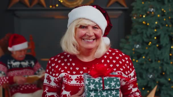 Seniorin im festlichen Pullover präsentiert Weihnachtsgeschenkschachtel, lächelt in die Kamera — Stockvideo