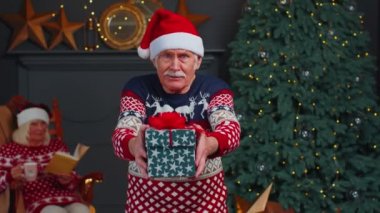 Yılbaşı süveteri ve şapka giymiş olgun büyükbaba Noel hediye kutusunu sunuyor, tatillerde evde.