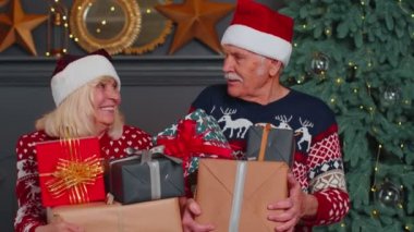 Noel Baba şapkalı yaşlı çift Noel hediyesi kutularını tutarken kameraya bakıyor.