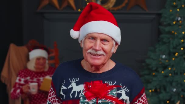 Senior nagyapa ünnepi pulóverben bemutatja karácsonyi ajándék doboz, mosolyog, belenéz a kamerába — Stock videók