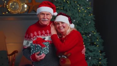 Noel 'i kutlamak için Noel Baba şapkalı yaşlı çift kameraya bakıyor ve evde birbirlerine sarılıyorlar.