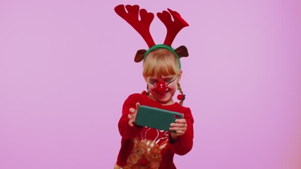 Νήπιο κορίτσι σε χριστουγεννιάτικα ελάφια κέρατα με ενθουσιασμό παίζει αγωνιστικά βιντεοπαιχνίδια στο κινητό τηλέφωνο — Αρχείο Βίντεο