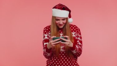 Endişeli kız Noel süveteri giyiyor, şapkası hevesle cep telefonuyla video oyunu oynuyor.