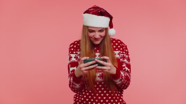 Ανήσυχη κοπέλα σε χριστουγεννιάτικο πουλόβερ, καπέλο ενθουσιωδώς παίζει αγωνιστικά βιντεοπαιχνίδια στο κινητό τηλέφωνο — Αρχείο Βίντεο
