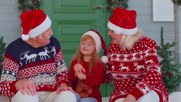 Abuelos mayores con nieta en el sombrero de Santa Claus celebrando la Navidad cerca de la casa decorada — Vídeos de Stock