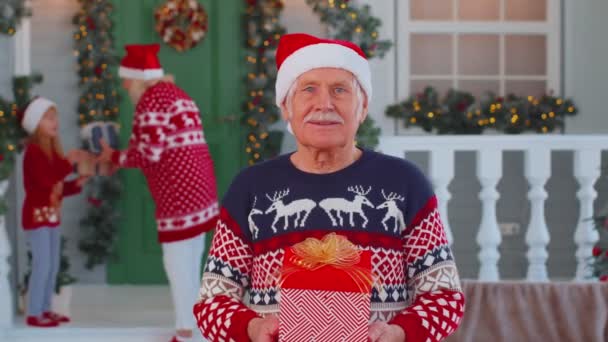 Portrét dědečka muž prezentuje dárkové krabice usmívá v blízkosti zdobené vánoční dům s rodinou — Stock video