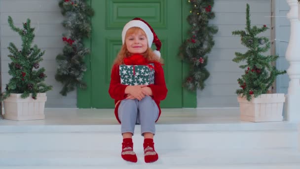 Joyeux souriant tout-petit enfant fille enfant assis à la maison décorée porche tenant une boîte de Noël — Video