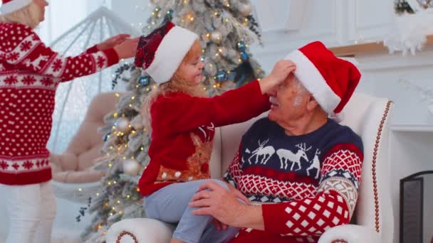 Wnuczka dziecko naprawia Boże Narodzenie Santa Claus kapelusz na szczęśliwy starszy stary uśmiechnięty dziadek w domu — Wideo stockowe