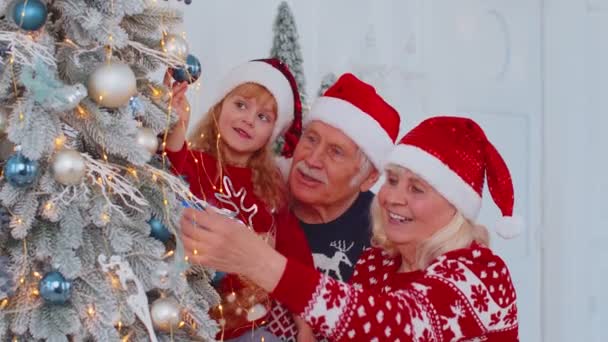 Niña con abuelos mayores decorando pino artificial de Navidad en la habitación de casa — Vídeos de Stock