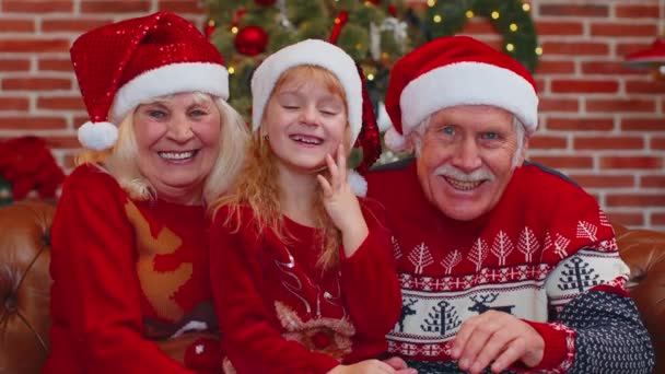 Abuelos mayores con nieta en sombreros de Papá Noel riendo en voz alta en casa cerca del árbol de Navidad — Vídeos de Stock