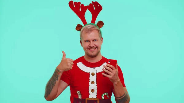 Чоловік у різдвяній футболці, використовуючи мобільний телефон, вводячи новий пост в Інтернеті, купуючи онлайн, показуючи великі пальці вгору — стокове фото