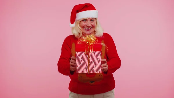 Зріла бабуся в новорічному светрі і капелюсі представляє різдвяну подарункову коробку, торговельні свята — стокове фото