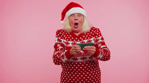 Беспокойная бабушка в рождественском свитере с энтузиазмом играет в гоночные видеоигры на мобильном телефоне — стоковое фото