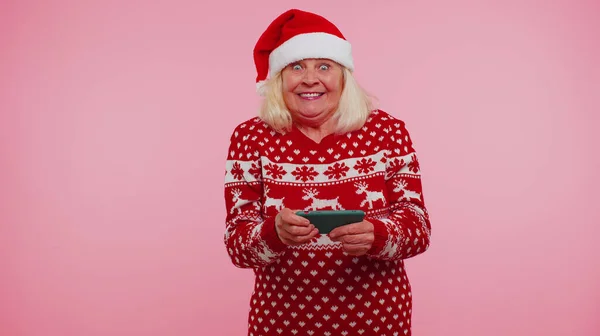 クリスマスセーター姿のおばあちゃんスマートフォンディスプレイ誠実に勝利成功運を喜ぶ — ストック写真