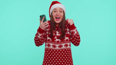 Young teen girl in Christmas sweater looking smartphone display sincerely rejoicing win success luck clipart