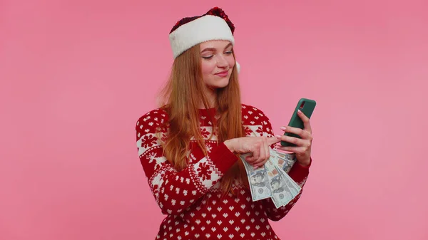 Ragazza in maglione rosso di Natale utilizzando il telefono cellulare, tenere soldi in contanti dollaro, facendo shopping online regalo — Foto Stock