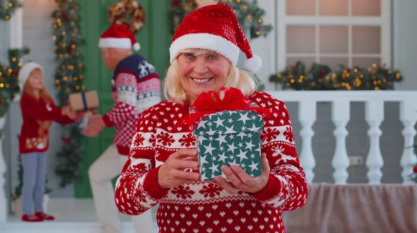 Portret babci kobieta prezentuje pudełko z uśmiechem obok ozdobionego domu Bożego Narodzenia z rodziną — Zdjęcie stockowe