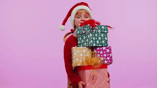 Menina em camisola de Natal, chapéu de Papai Noel, sorrindo, segurando muitas caixas de presente — Fotografia de Stock