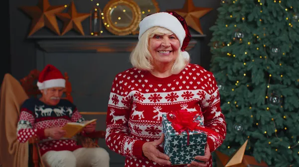 Старша бабуся в святковому светрі представляє різдвяну подарункову коробку, посміхаючись, дивлячись на камеру — стокове фото
