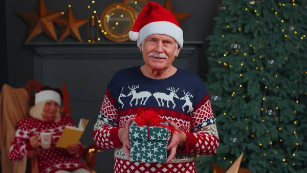 Älterer Großvater im festlichen Pullover präsentiert Weihnachtsgeschenkbox, lächelt, blickt in die Kamera — Stockfoto