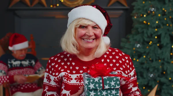 Старшая бабушка в праздничном свитере представляет рождественскую подарочную коробку, улыбается, смотрит в камеру — стоковое фото