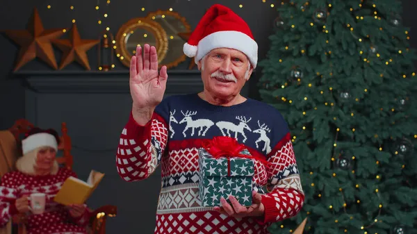 Senior grandfather in Christmas sweater smiling friendly at camera and waving hands gesturing hello — ストック写真