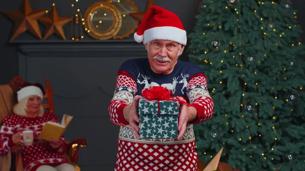 Зрілий дідусь в новорічному светрі і капелюсі представляє різдвяну подарункову коробку, свята вдома — стокове фото