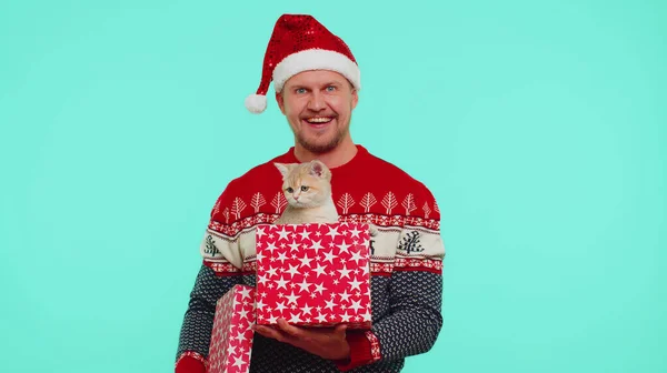 Чоловік у різдвяному червоному капелюсі светр посміхається, розгорнувши подарунок, відкриваючи коробку з домашнім котом, чудовий сюрприз — стокове фото