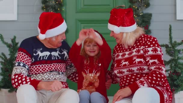 Elderly grandmother grandfather with granddaughter sitting at Christmas house porch waving hello hi — Stock Video