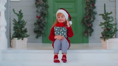 Neşeli gülümseyen küçük kız çocuk elinde bir Noel kutusuyla verandada oturuyor.