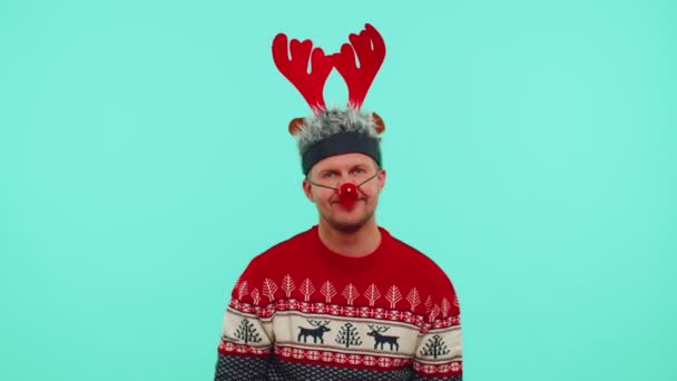 Веселий милий 30-річний чоловік носить червоний новорічний светр і капелюх посміхається, дивлячись на камеру — стокове відео
