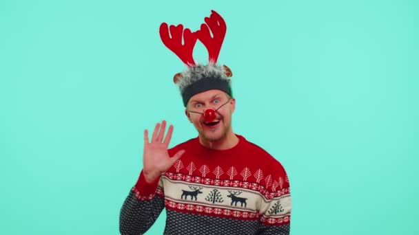 Hombre positivo en suéter de Navidad, cornamenta de ciervo olas palma de la mano en gesto de hola da la bienvenida a alguien — Vídeos de Stock