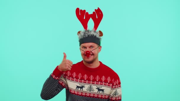 Funny man wears red New Year sweater deer antlers raises thumbs up agrees with something good, like — Stock Video