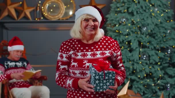 Eldre bestemor i festlig genser som presenterer julegave, smiler, ser på kamera – stockvideo