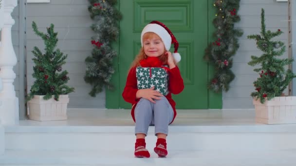 Fröhlich lächelnde Kleinkind Kind Mädchen Kind sitzt am geschmückten Haus Veranda hält eine Weihnachtsschachtel — Stockvideo