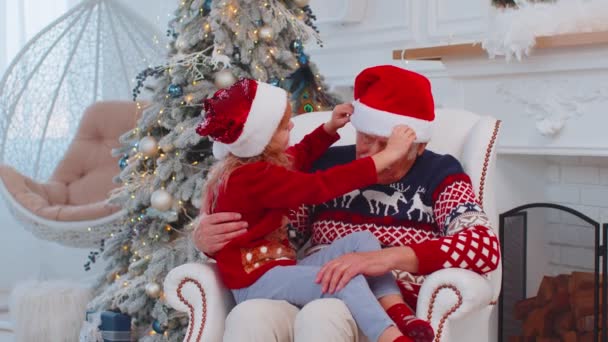 Девочка-внучка чинит рождественскую шляпу Санта-Клауса на счастливом старом улыбающемся дедушке дома — стоковое видео