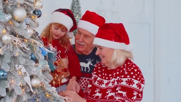 Счастливые старые бабушка и дедушка, внучка малыша висит игрушки украшая елку празднуя — стоковое видео