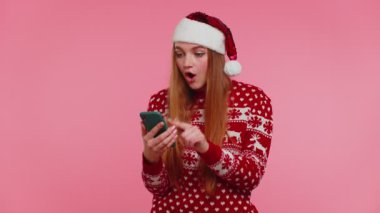 Şık bir Noel kazağı giyen, akıllı telefon görünümlü, neşeli, başarılı bir zafer kazanan kadın.