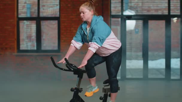 ヘイズジムでの静止自転車トレーニングルーチンの回転に乗るアスレチック女性、体重減少室内 — ストック動画
