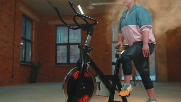 Atletická dívka provádějící aerobní jízdu na koni cvičení na kole stacionární kolo v mlhavé tělocvičně — Stock video