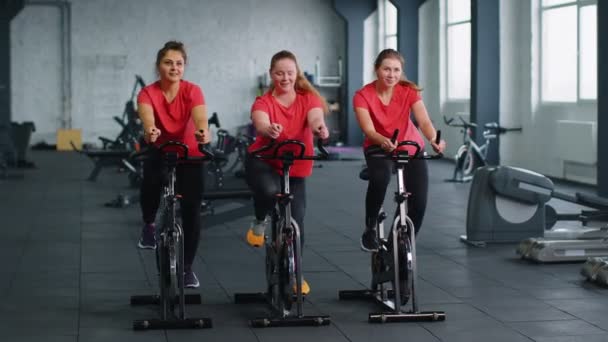 Υγιής Καυκάσιος ομάδα γυναικών άσκηση προπόνηση σε σταθερό ποδήλατο μηχανή ποδήλατο στο γυμναστήριο — Αρχείο Βίντεο