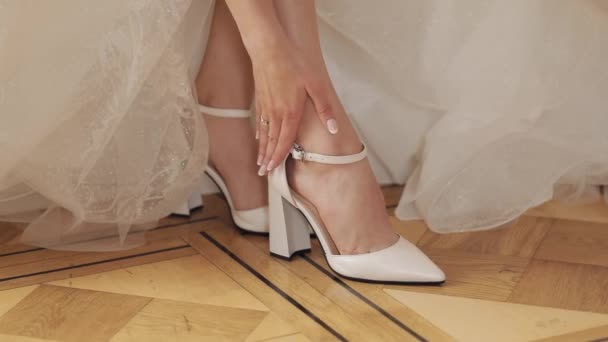 Junge Braut im weißen Hochzeitskleid trägt hochhackige Schuhe an schlanken Beinen, während sie zu Hause sitzt — Stockvideo