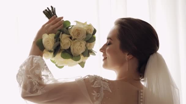 Νύφη σε λευκό φόρεμα που διαμένουν κοντά στο παράθυρο με λουλούδια μπουκέτο, γάμο πρωινές προετοιμασίες στο σπίτι — Αρχείο Βίντεο