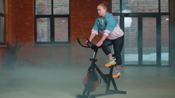 Healthy Caucasian woman exercising workout on stationary cycling machine bike in gym, slow motion — Stock Video